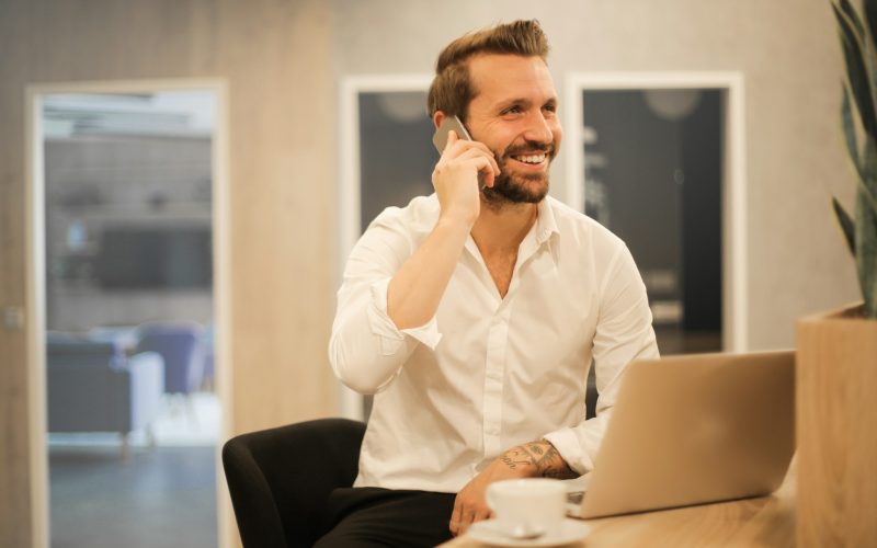 en leende man på telefon bakom sin laptop, glad över sing framgångsrika ekonomiska planering